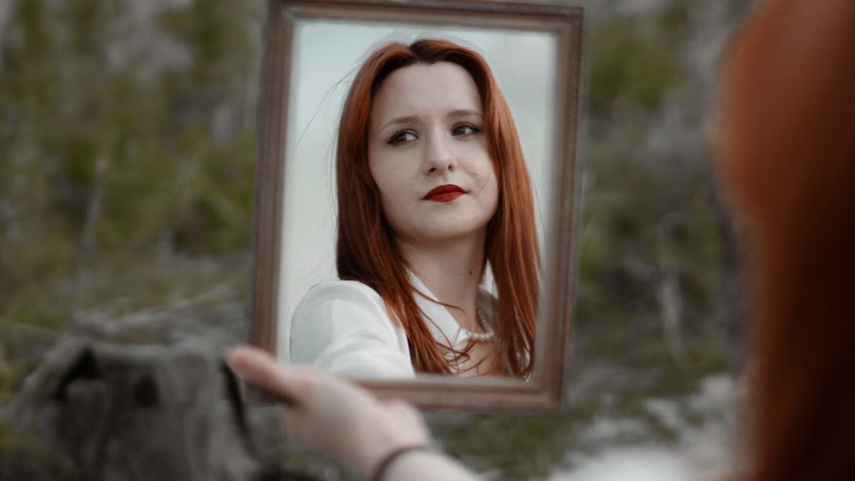 A young redhead woman holding a mirror outdoors, showcasing her reflection with red lips.